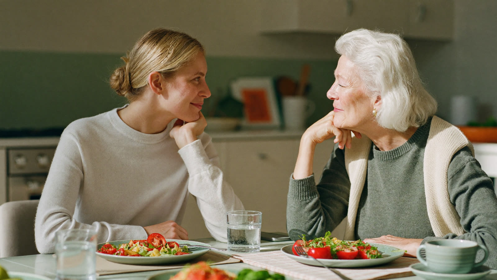 An older woman and her adult daughter talking together at a sunlit kitchen table
