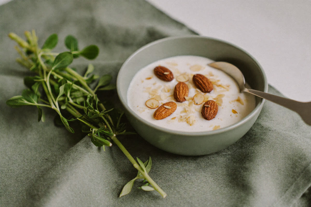 A bowl of yogurt topped with almonds beside fresh greens on a linen cloth