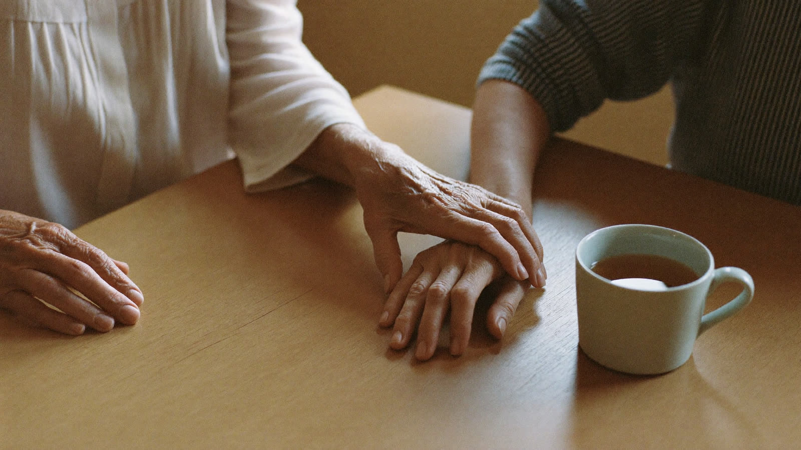 An older woman's hand resting over another's across a wooden table beside a cup of tea