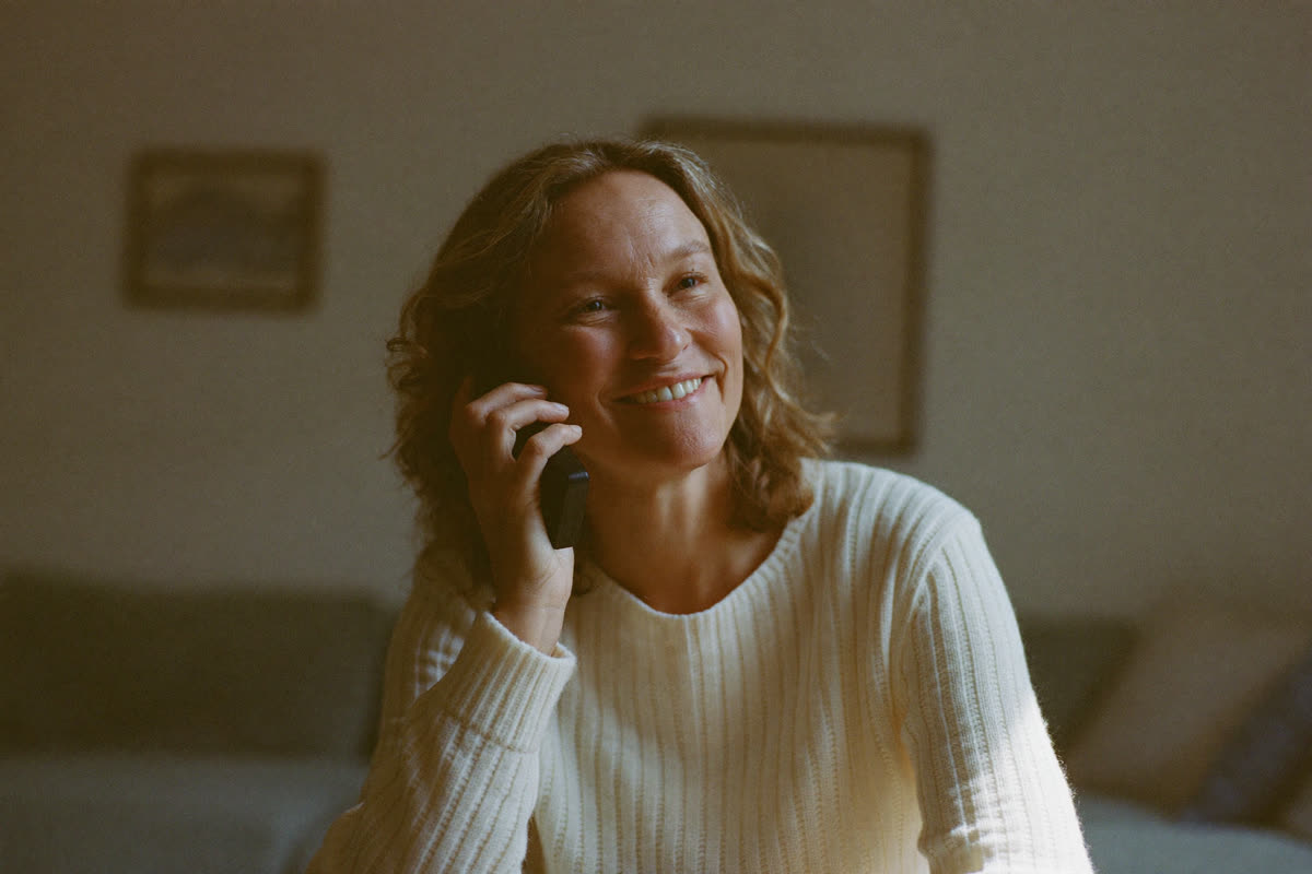 Woman in a cream sweater smiling as she listens on the phone in warm window light