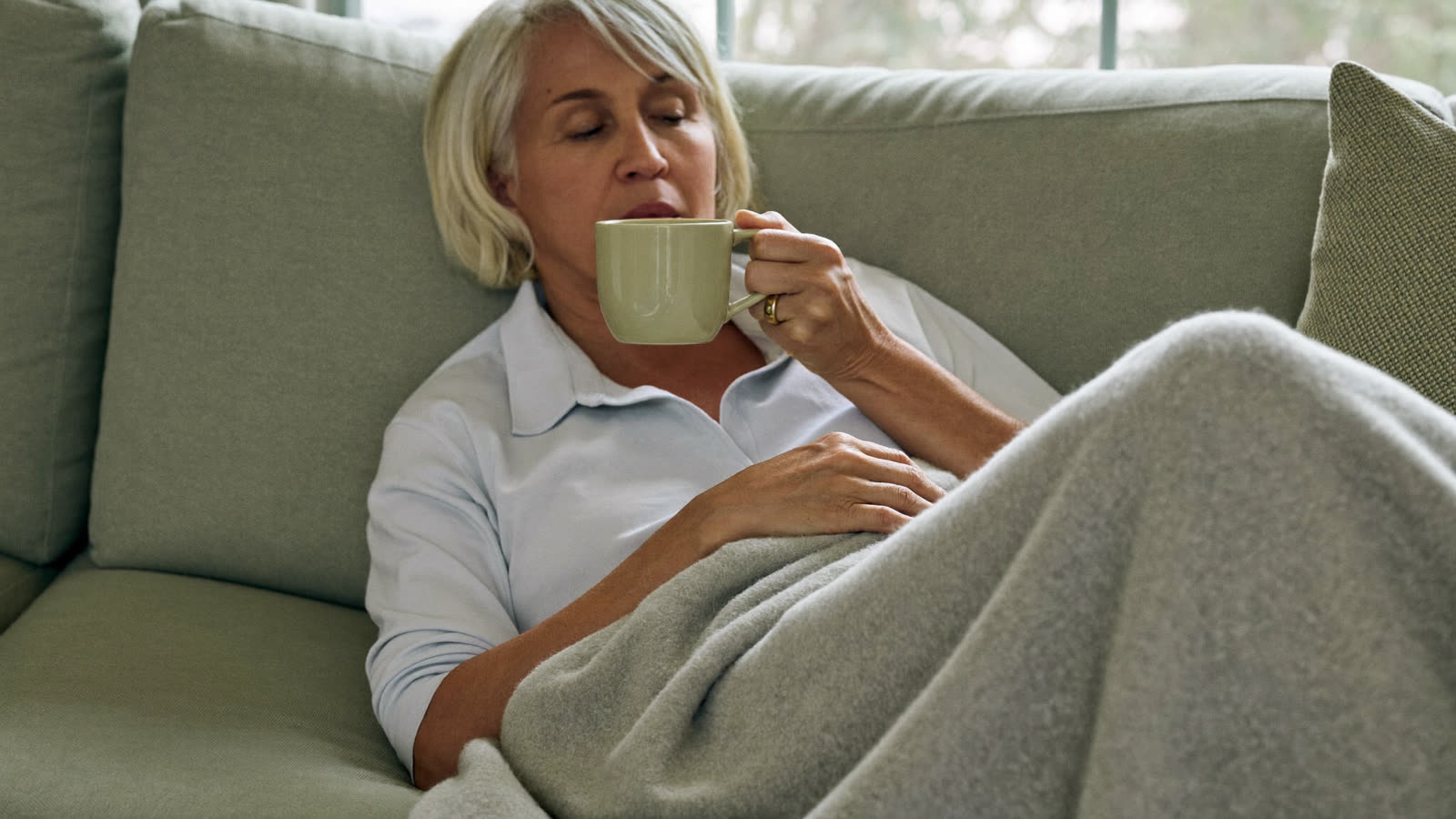 Woman in her forties wrapped in a blanket on a sofa holding a warm mug