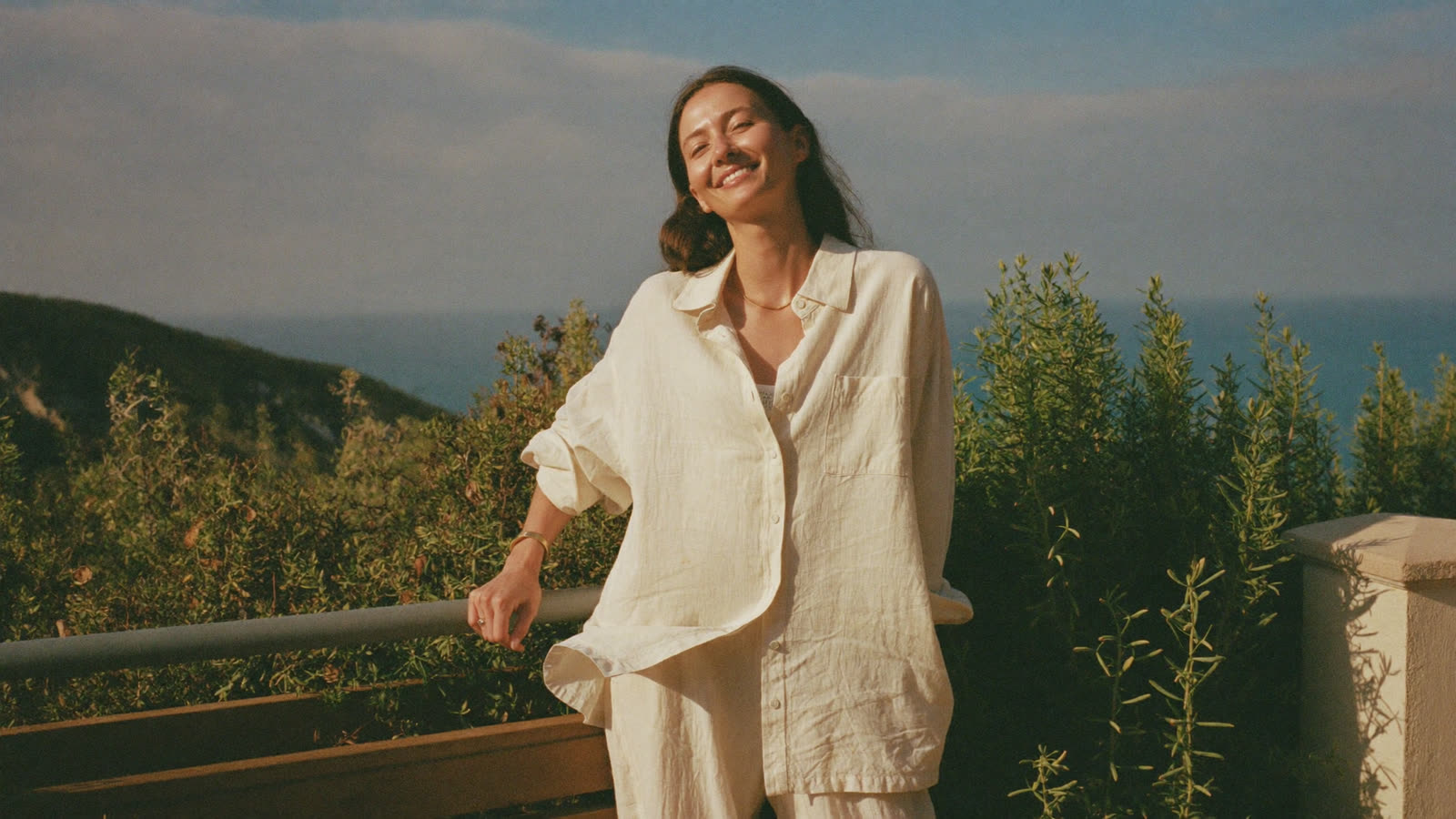 Relaxed, smiling woman in her sixties on a sunlit coastal terrace in golden light