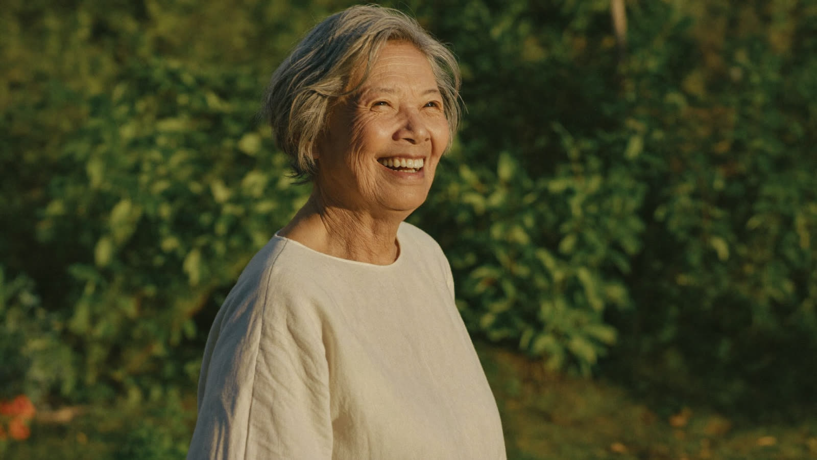 Woman in her seventies smiling warmly outdoors in a sunlit garden